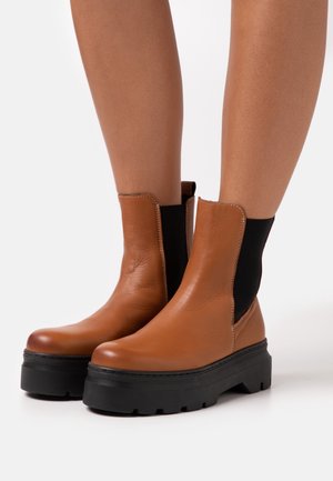 Stivaletti alla caviglia in pelle marrone con pannelli laterali elastici neri e spesse suole platform nere indossati su gambe nude.