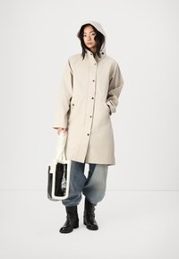 Beige regnrock med huva, tryckknappar, sidofickor, ihop med blå vida jeans och svarta ankelskots. Tillbehören inkluderar en svart och vit tote.