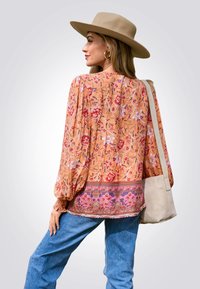 Langärmlige Blumenbluse in Orange mit einem gemusterten Saum und elastischen Bündchen, kombiniert mit hellblauen Jeans und einer beigen Schultertasche.