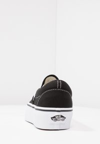 Zapato sin cordones de lona negra con costuras blancas y una suela blanca gruesa. Presenta una etiqueta de logo en el talón y un diseño en blanco y negro contrastante.