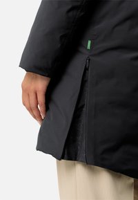 Giacca impermeabile nera con zip laterale, caratterizzata da una superficie matta e liscia e un'etichetta verde discreta, contrastata da un tessuto beige sottostante.