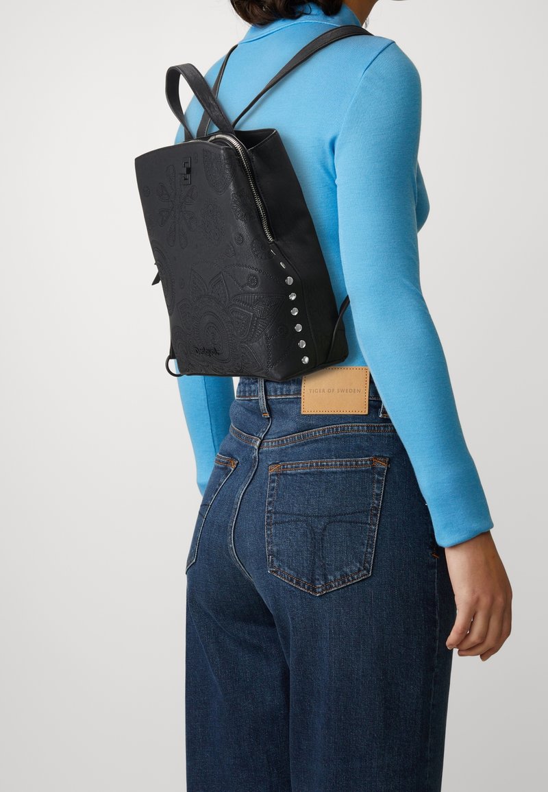 Czarny skórzany plecak z tłoczonymi kwiatowymi wzorami, srebrnymi ćwiekami po bokach i zamkiem na górze. Nosi się go na plecach, na niebieskiej bluzce.