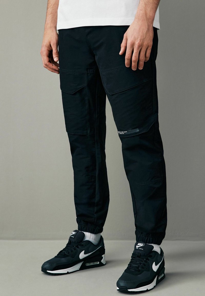 Sorte cargo bukser med elastiske manchetter, flere lommer og en glat tekstur, parret med sort og hvid Nike Air Max sneakers.
