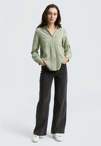 Camicia verde chiaro con bottoni e superficie strutturata, con colletto e due tasche sul petto, abbinata a pantaloni neri a zampa e sneakers bianche.