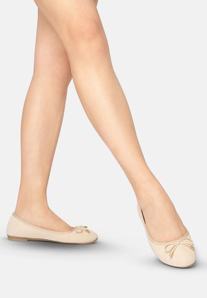 Beine gekreuzt, beige Ballerinas mit kleinem Schleifchen und goldenen Spitzen auf hellgrauem Hintergrund.