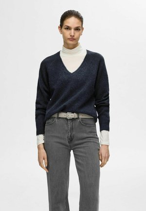 Svetřík s námořnickým výstřihem a dlouhými rukávy je přehozený přes bílý rolák, v kombinaci s šedými džíny s vysokým pasem a světle šedým opaskem.