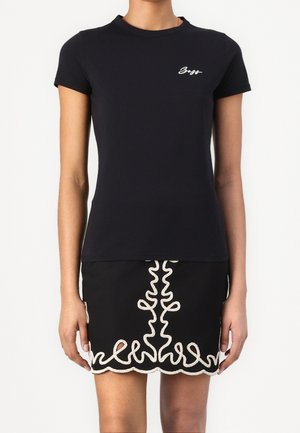 Vrouw draagt een nauwsluitend zwart T-shirt met korte mouwen met wit "Boss"-logo en een zwarte rok met witte geborduurde geschulpte zoom.