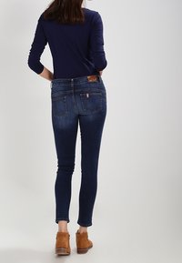 Jeans de mezclilla azul ajustados con una cintura alta, con bolsillos traseros y una etiqueta de cuero. Combinados con botas cortas de color marrón claro y una parte superior oscura.