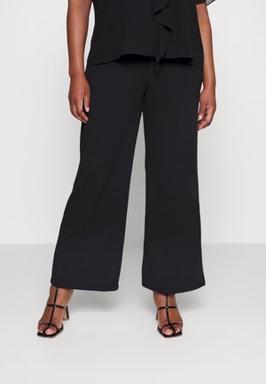 Pantalon large noir porté avec des sandales à talons ouvertes noires, présenté sur une personne debout devant un fond uni.