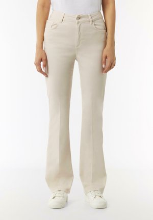 Beige Schlagjeans aus einem weichen, glatten Material mit Fronttaschen und einem Knopfverschluss, kombiniert mit weißen Sneakers.