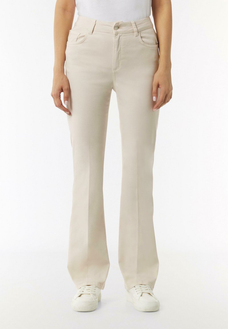 Beige Schlagjeans aus einem weichen, glatten Material mit Fronttaschen und einem Knopfverschluss, kombiniert mit weißen Sneakers.