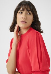 Pull en tricot rouge avec des manches courtes bouffantes, un col rond et une texture lisse. Le mannequin touche son cou, tourné légèrement de profil par rapport à la caméra.