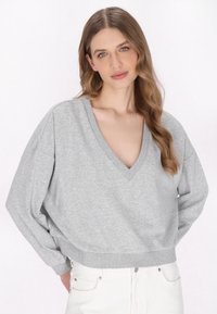 Grauer Pullover mit V-Ausschnitt, lässigem Schnitt und geripptem Saum. Weicher Stoff mit überschnittenen Schultern, kombiniert mit weißen Jeans.
