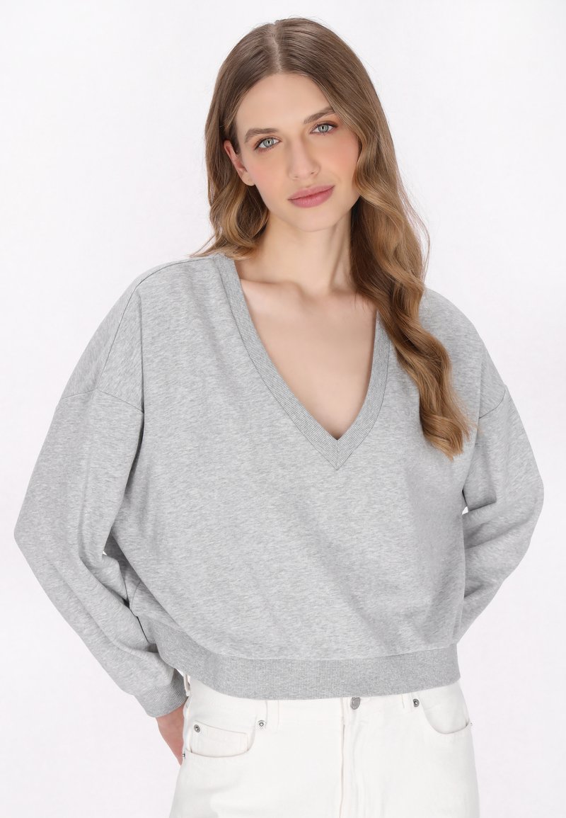 Grauer Pullover mit V-Ausschnitt, lässigem Schnitt und geripptem Saum. Weicher Stoff mit überschnittenen Schultern, kombiniert mit weißen Jeans.
