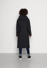 Abrigo acolchado negro con capucha, con líneas acolchadas horizontales, mangas largas y longitud por debajo de la rodilla, usado sobre jeans oscuros y zapatillas.