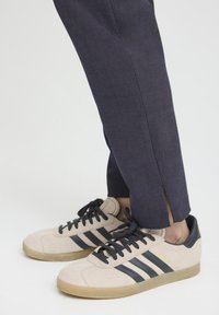 Suede beige sneakers with black stripes, black laces, and a gum sole. Paired with dark blue pants featuring side slits.