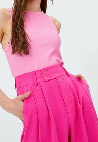 Różowy prążkowany top na ramiączkach w połączeniu z różowymi spodniami z wysokim stanem i plisami, które mają dwa widoczne guziki i kieszenie po bokach. Gładka faktura.