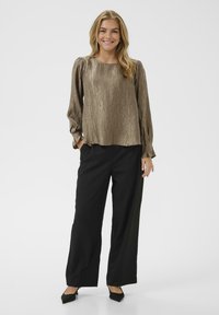 Donna in piedi che sorride, indossando una blusa lunga a maniche lucide di bronzo, pantaloni neri a gamba larga e décolleté nere a punta.