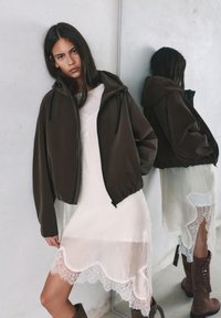 Tânără îmbrăcată într-o rochie albă din dantelă poartă o jachetă supradimensionată de culoare închisă și cizme maro cu șireturi, sprijinindu-se de o oglindă pe un fundal de perete deschis la culoare.