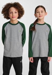Grijze longsleeve shirts met groene raglanmouwen, met een klein logo op de borst. Beide zijn gecombineerd met zwarte broeken.