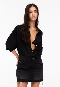Chemise noire oversize à boutons, manches retroussées, portée sur un bikini noir. Associée à une mini-jupe en denim noir avec un ourlet effiloché.