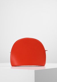 Czerwony skórzany worek w kształcie półokręgu z widocznym przeszyciem, umieszczony na białym cokole na tle jasnoszarym.