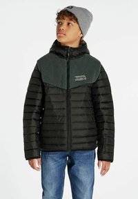 Giacca a vento nera con spalle in pile verde. Caratterizzata da chiusura con zip, maniche lunghe e una toppa con testo. Indossata con jeans blu e un berretto grigio.