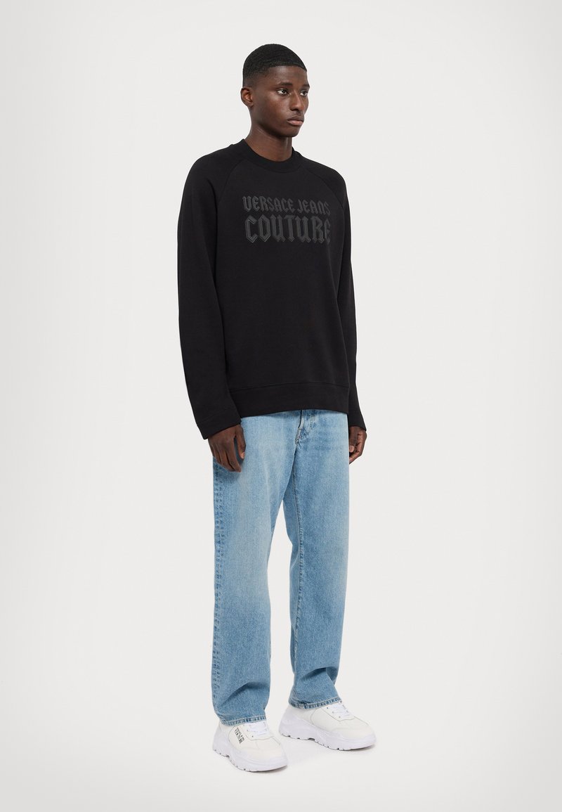 Černá mikina se vzorem tmavého loga "VERSACE JEANS COUTURE", doplněná světle modrými džíny s rovnými nohavicemi a bílými teniskami.
