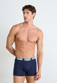 Temno modri bokserji iz mehkega materiala z zeleno-belo elastično pasom, na katerem je vidno prikazan logotip "PUMA".