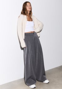 Hellbeige Cardigan, weißes kurzes Tanktop, graue plissierte Hosen mit weitem Bein und weiße Turnschuhe. Glatter Stoff mit lockerer Passform und dezentem Fall.