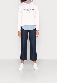 Felpa rosa con logo "TOMMY HILFIGER", indossata sopra una camicia a righe blu e bianche, abbinata a jeans svasati scuri e scarpe da ginnastica bianche.