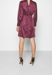 Robe fuchsia à manches longues, avec un dos en V et un motif floral. Le tissu est d'une texture lisse. Associée à des chaussures à talons hauts noires.
