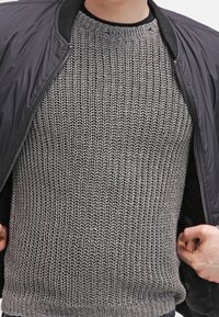 Maglione grigio a maglia con un motivo testurizzato, indossato sotto una giacca bomber nera, che mette in evidenza polsini a coste e una scollatura ampia.