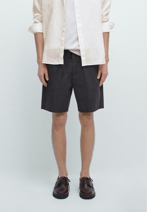 Person trägt beige langärmliges Hemd, weißes T-Shirt, schwarze knielange Shorts und dunkelbraune Schnürschuhe, steht vor hellem einfarbigem Hintergrund.