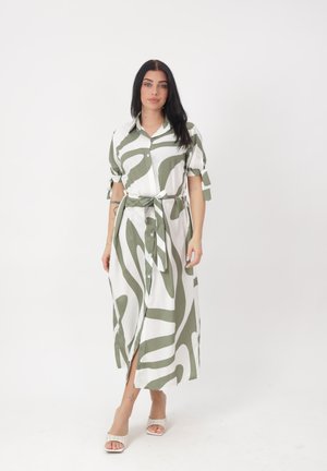 Kobieta o długich ciemnych włosach, ubrana w białą sukienkę z oliwkowozielonym abstrakcyjnym nadrukiem, zapinaną na guziki, z krótkimi rękawami i paskiem związanym w talii, idąca do przodu.