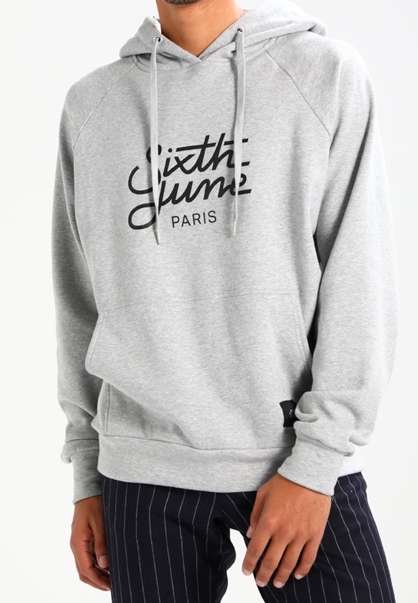 Homme portant un sweat à capuche gris clair avec le texte "Sixth June PARIS", associé à un pantalon sombre à fines rayures, debout dans une posture détendue.