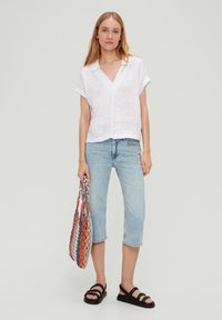 Biała lniana koszula z krótkim rękawem, dekolt w serek, w zestawieniu z jasnoniebieskimi, ściętymi dżinsami i czarnymi sandałami. Trzyma wielokolorową plecioną torbę.