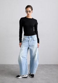 Maglietta nera a maniche lunghe, jeans a palazzo blu chiaro e sneakers nere. Il modello indossa occhiali da sole grigi; l'outfit presenta una vestibilità rilassata e orli sfrangiati.
