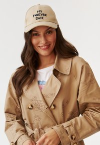 Beige trench coat illusztrált mintákkal és arany gombokkal, bézs sapkával, amelyen fekete hímzett szöveg található. Alatta fehér póló.