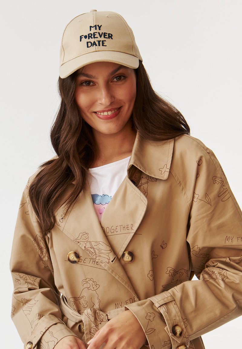 Beige trench coat illusztrált mintákkal és arany gombokkal, bézs sapkával, amelyen fekete hímzett szöveg található. Alatta fehér póló.