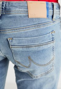 Vaaleansiniset farkut, joissa on vaaleanruskea nahkainen merkki takavyötäröllä, näkyvät saumat ja kaksi takataskua, joissa on kaarevat yksityiskohdat.