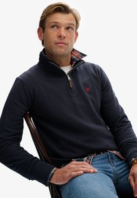 Navyblauwe zip-up sweatshirt met rood logo, met een geruite binnenkraag en manchetten. Gestyled met blauwe jeans, zittend op een houten stoel.