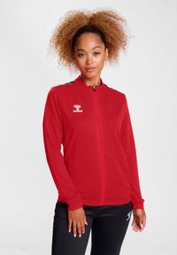 Rote Zip-Jacke aus weichem Material, mit langen Ärmeln, hohem Kragen und schwarzen Akzenten an den Schultern. Hummel-Logo auf der Brust.