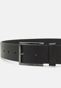 Curea din piele neagră, cu o cataramă dreptunghiulară din argint care prezintă logo-ul brandului. Textură netedă, mai multe orificii de ajustare și un design elegant.