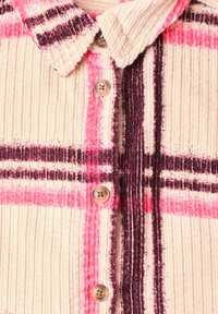 Gros plan d'une chemise en tissu côtelé crème avec des rayures à carreaux roses et violet foncé et des boutons beige sur le devant.