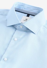 Himmelblaues strukturiertes Hemd mit einem formellen Kragen, ausgestattet mit weißen Knöpfen und einem gemusterten Futter unter dem Kragen. Markenlabel sichtbar.