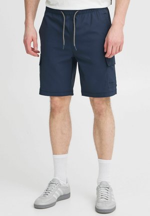 Shorts cargo blu navy con vita regolabile tramite coulisse, tasche laterali e una texture liscia. Presenta un piccolo logo sulla gamba sinistra.