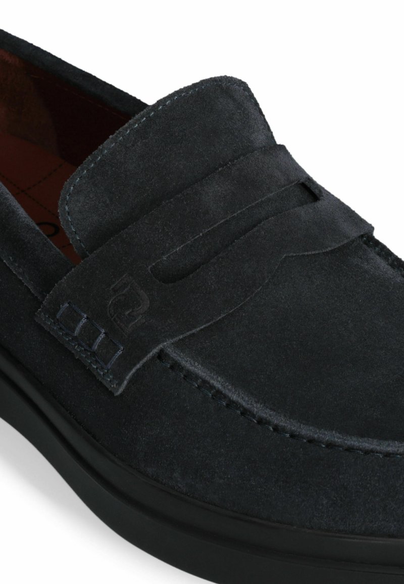 Zapato negro de ante tipo slip-on con puntera redondeada, correa decorativa y detalles texturizados. Cuenta con un forro interior suave y una suela de goma negra duradera.