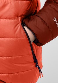 Padded orange Jacke mit Seitentasche. Glatte Textur, kontrastierender brauner Ärmel mit Logodetail. Der Reißverschluss hat einen blauen Ziehgriff.