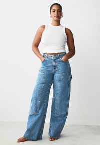 Jeans a vita alta in denim blu a gamba larga, con grandi tasche frontali e orlo sfrangiato, abbinati a un top aderente bianco.